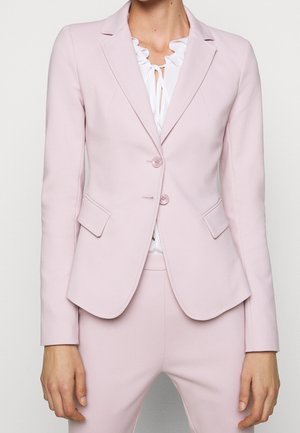 Světle růžový blazer s přizpůsobeným střihem, klopami, dvěma knoflíky a bočními kapsami. V kombinaci s bílu blůzou s volánky pod ním.