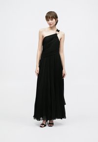 Fekete pliszírozott maxiruha aszimmetrikus nyakkivágással, egypántos részlettel és légies textúrával. Fekete magas sarkú szandállal viselve.