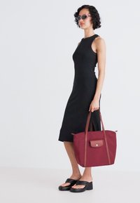 Black sleeveless dress with a side slit, paired with a burgundy tote bag featuring brown leather accents and two shoulder straps.