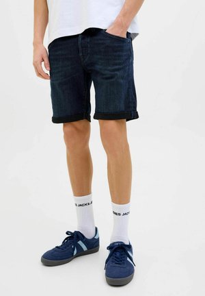 Donkerblauwe denimshorts met omgeslagen mouwen, gecombineerd met witte sokken met zwarte tekst en blauwe sneakers. De shorts hebben voorzakken.