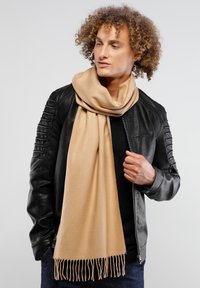 Bufanda beige hecha de un tejido suave, con flecos en los bordes, caída de forma holgada alrededor del cuello sobre una chaqueta negra.