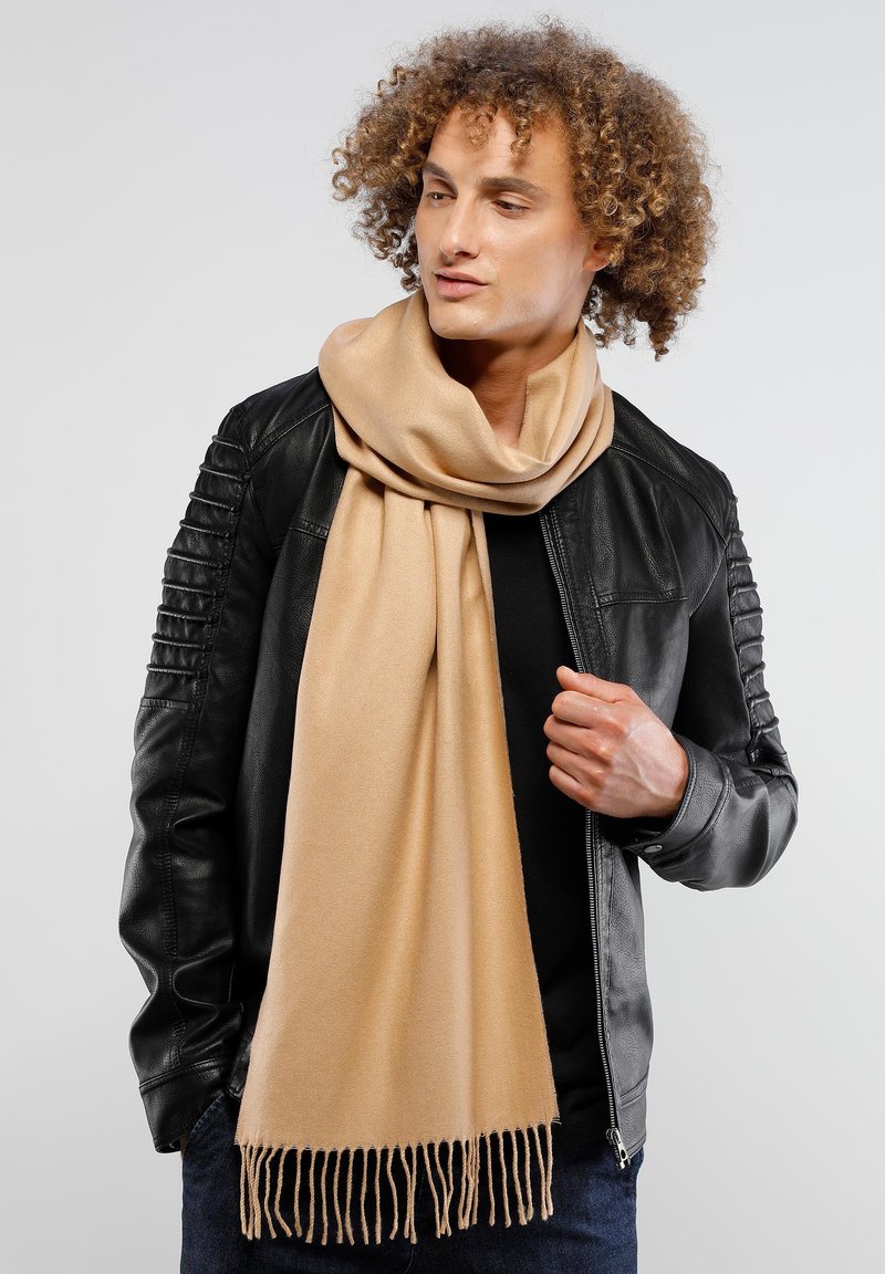 Bufanda beige hecha de un tejido suave, con flecos en los bordes, caída de forma holgada alrededor del cuello sobre una chaqueta negra.
