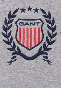 Tela gris con un escudo bordado que tiene franjas rojas, hojas de laurel en azul marino y la palabra "GANT" arriba, acentuado por tres estrellas encima del escudo.