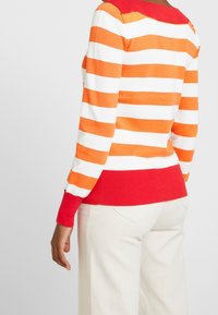 Pull rayé orange et blanc avec un col et un ourlet côtelés rouges. Il présente des manches longues et une coupe ajustée.