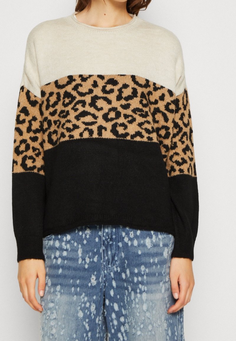 Femme portant un pull en bloc de couleurs avec des sections beige, imprimé léopard et noir, associé à un jean bleu délavé à l'acide.