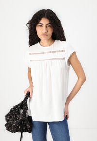 White short-sleeve top with lace detailing across the chest, round neckline, and a relaxed fit. Paired with a black floral bag.