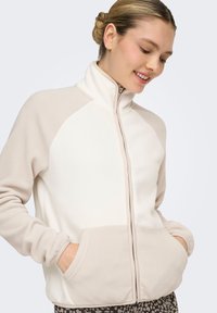 Veste zippée en polaire crème et beige, dotée de manches raglan, de poches latérales et d'un col montant. Texture douce avec un design bicolore.
