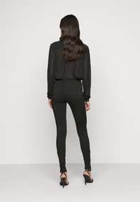 Blusa negra transparente de manga larga combinada con jeans negros ajustados, destacando una textura suave y una silueta entallada. Sandalias de tacón alto incluidas.