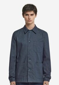 Mörk denimjacka med krage, knäppning framtill med knappar och två fickor på framsidan. Har blåa knappar och ett lätt texturerat tyg.