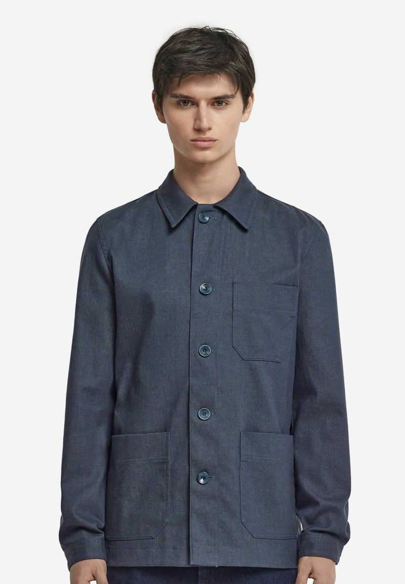 Mörk denimjacka med krage, knäppning framtill med knappar och två fickor på framsidan. Har blåa knappar och ett lätt texturerat tyg.