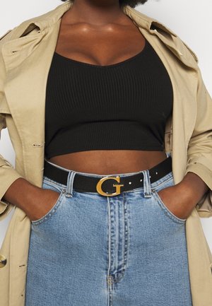 Kobieta w beżowej kurtce, czarnym krótkim prążkowanym topie, niebieskiej spódnicy jeansowej z wysokim stanem oraz czarnym pasku z złotą klamrą w kształcie litery "G".