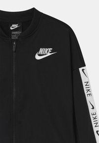 Chaqueta negra de Nike con cremallera frontal, cuello acanalado y logo blanco. Presenta una franja blanca en la manga con el texto "Nike" repetido. Textura suave.