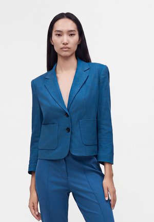 Femme aux longs cheveux noirs portant un blazer bleu ajusté et un pantalon assorti, debout avec une main sur la hanche et des talons beiges.