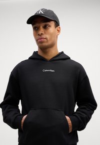 Črna kapuca s sprednjim žepom, s belim logotipom "Calvin Klein". V kompletu s črnimi kapicami z vezenim črko "K".