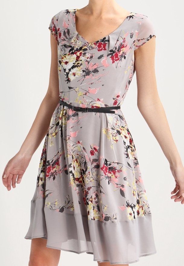 Femme portant une robe fleurie gris clair avec des fleurs roses, rouges, jaunes et noires, des manches courtes et une fine ceinture noire à la taille.