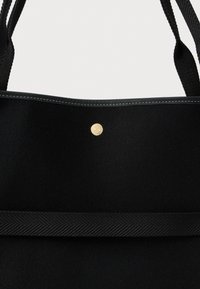 Μαύρη τσάντα tote με υφασμάτινη οριζόντια ταινία κοντά στη βάση, μαύρους ραμμένους χειροκίνητους και μία στρογγυλή χρυσή κουμπί κλεισίματος στο μπροστινό μέρος.