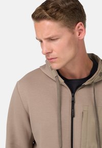 Khaki mikina s kapucňou a zipsom, vreckom na zips a kontrastnými textúrovanými akcentmi. Hladký povrch a minimalistický dizajn.