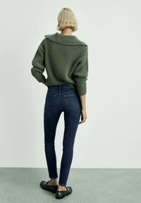 Maslinasto zeleni pleteni pulover s širokim ovratnikom, uparen s tamnoplavim uskim trapericama i crnim mokasinkama, prikazan s leđa.