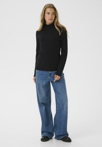 Zwarte geribbelde trui met col, gecombineerd met lichtblauwe wijde jeans en zwarte schoeisel. Model staat tegen een effen witte achtergrond.