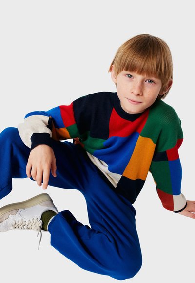 Suéter de punto multicolor con patrones rectangulares en rojo, azul, verde, negro y beige; combinado con pantalones azules y zapatillas blancas.