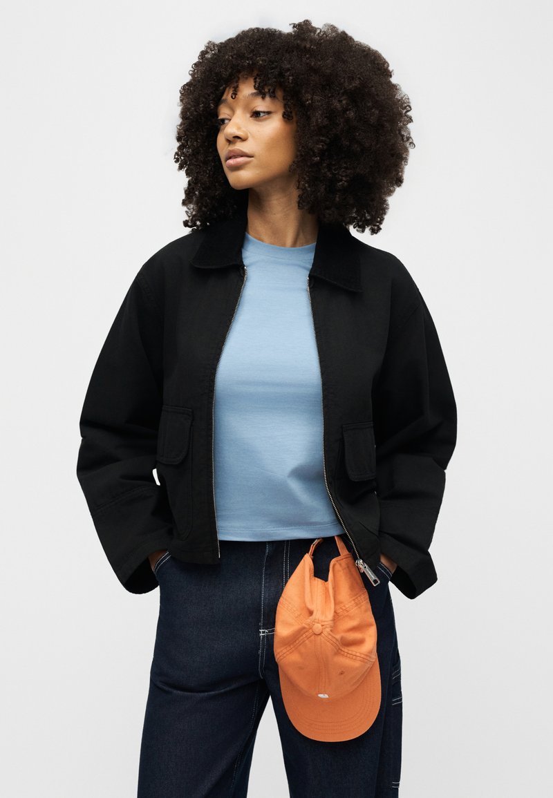 Woman with curly hair wearing black jacket, light blue shirt, dark jeans, hands in pockets, and orange cap hanging from waistband.