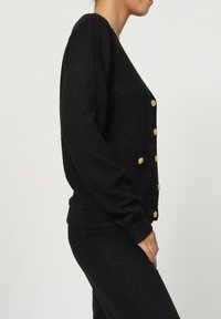 Cardigan noir à manches longues, avec des boutons dorés et deux poches avant. Matière en tricot texturé, design arrivant au genou.