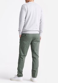 Man die met zijn rug naar de camera staat, gekleed in een lichtgrijze sweatshirt, olijfgroene broek met achterzakken, en witte sneakers, op een witte achtergrond.