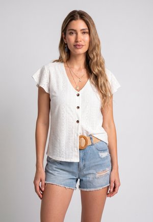 Frau mit langem welligem Haar, die eine weiße, geknöpfte Bluse, geschichtete goldene Halsketten, Jeansshorts mit Rissen und einen geflochtenen Gürtel mit runder Schnalle trägt.
