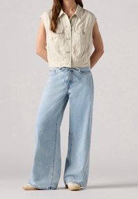 Ljust beige ärmlös denimjacka med knäppning och två framfickor, kombinerad med lösa ljusblå vida jeans.