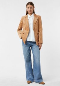 Marynarka sztruksowa w jasnobrązowym kolorze, zapinana na jeden guzik, z dwoma przednimi kieszeniami, w zestawie z białą koszulą i niebieskimi dżinsami o szerokich nogawkach.
