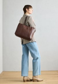 Brun læder tote taske med to håndtag, der har vertikal syning og et enkelt design. Modellen bærer lyseblå jeans og beige støvletter.