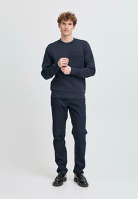 Marineblauwe sweatshirt met een borstzak, gedragen met donkere spijkerbroeken en zwarte schoenen. De stof lijkt glad en heeft een relaxte pasvorm.