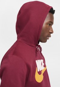 Hombre con sudadera burdeos y logo de Nike amarillo, mostrado de perfil mirando hacia la derecha contra un fondo liso.