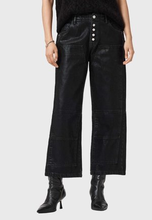 Vrouw draagt zwarte broek met wijde pijpen, grote voorzakken en zilveren knopen, gecombineerd met zwarte enkellaarzen met hak.