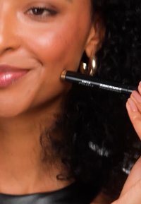 Bronze makeup applicator held near cheek, featuring a sleek black body with metallic accents. Curly hair and golden hoop earring visible.