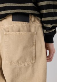 Pantalons beiges avec une poche arrière, texture denim, et une étiquette noire sur la taille. La personne porte un pull rayé noir et vert.