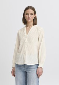 Blusa di colore crema chiaro con righe verticali, scollatura a V, chiusura con bottoni e maniche lunghe. Tessuto morbido e leggero con una vestibilità comoda.