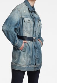 Džínová bunda ve vybledlých modrých tónech, s páskem v pase, velkými kapsami na hrudi a zapínáním na knoflíky s viditelnými detaily šití.