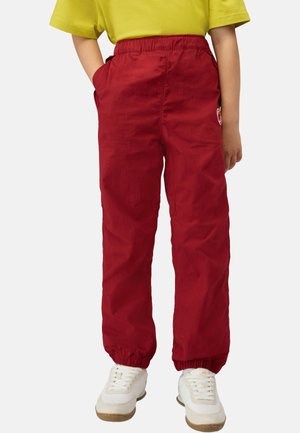 Barn iført røde elastik-bukser med elastiske manchetter, gul top, hvide sneakers, stående med hænderne i lommerne på en ensfarvet baggrund.