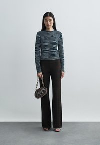 Top de malha às riscas preto e branco com mangas longas, emparelhado com calças flare texturizadas escuras. O modelo segura uma pequena bolsa estampada com alça de corrente.