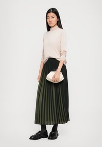 Light pink, long-sleeve turtleneck paired with a pleated black and dark green midi skirt. Model holds a white clutch and wears black loafers.