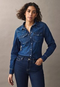Cămașă din denim de culoare albastru închis, cu nasturi în față, două buzunare pe piept, coasere albe și mâneci lungi. Asociată cu blugi negri.