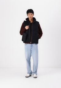 Zwarte puffer vest met een rits aan de voorkant, gedragen over een bruine trui. Lichte blauwe losse jeans en een zwarte crossbody tas. Witte en grijze sneakers.
