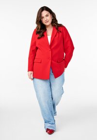 Roter Blazer mit einem einzigen Knopf, Reverskragen und Fronttaschen, kombiniert mit hellblauen, weiten Jeans und roten Schuhen. Glatter Stoff.