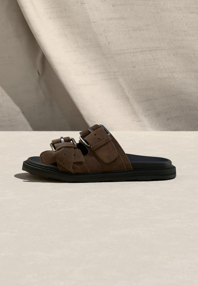 Καφέ σανδάλι slide από σουέντ με δύο ρυθμιζόμενους ιμάντες με αγκράφες και μαύρη σόλα, σε υπόλευκο φόντο με υφή.