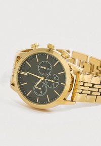 Kronografski ručni sat zlatne boje s crnim brojčanikom, tri podbrojčanika, pokazivačima sati u obliku štapića i metalnom narukvicom od karika. Glatka tekstura završne obrade.