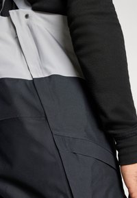 Giacca a strati nera e grigia con un design elegante, tessuto texturizzato, chiusura a scatto e accenti minimalisti.