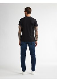 Černé tričko s krátkým rukávem a kulatým výstřihem, oblečené s tmavě modrými džíny a bílými teniskami, které ukazuje detailní tetování na pažích.