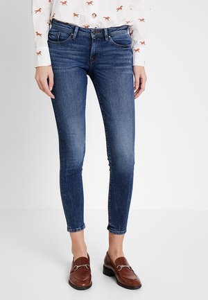 Femme portant un jean skinny bleu foncé, une chemise blanche avec des imprimés de chevaux marron et des mocassins en cuir marron, debout devant un fond blanc.