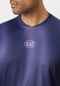Mornarsko modra športna majica z V-izrezom, izdelana iz gladkega materiala, z belim logotipom EA7 na prsih.